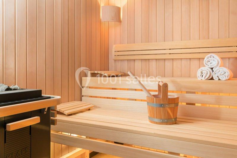 Bancs en bois clair dans un sauna avec seau, louche et serviettes roulées posés sur une étagère.