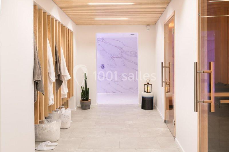 Couloir lumineux avec serviettes suspendues, plantes, et accès à une douche en marbre et un sauna vitré.