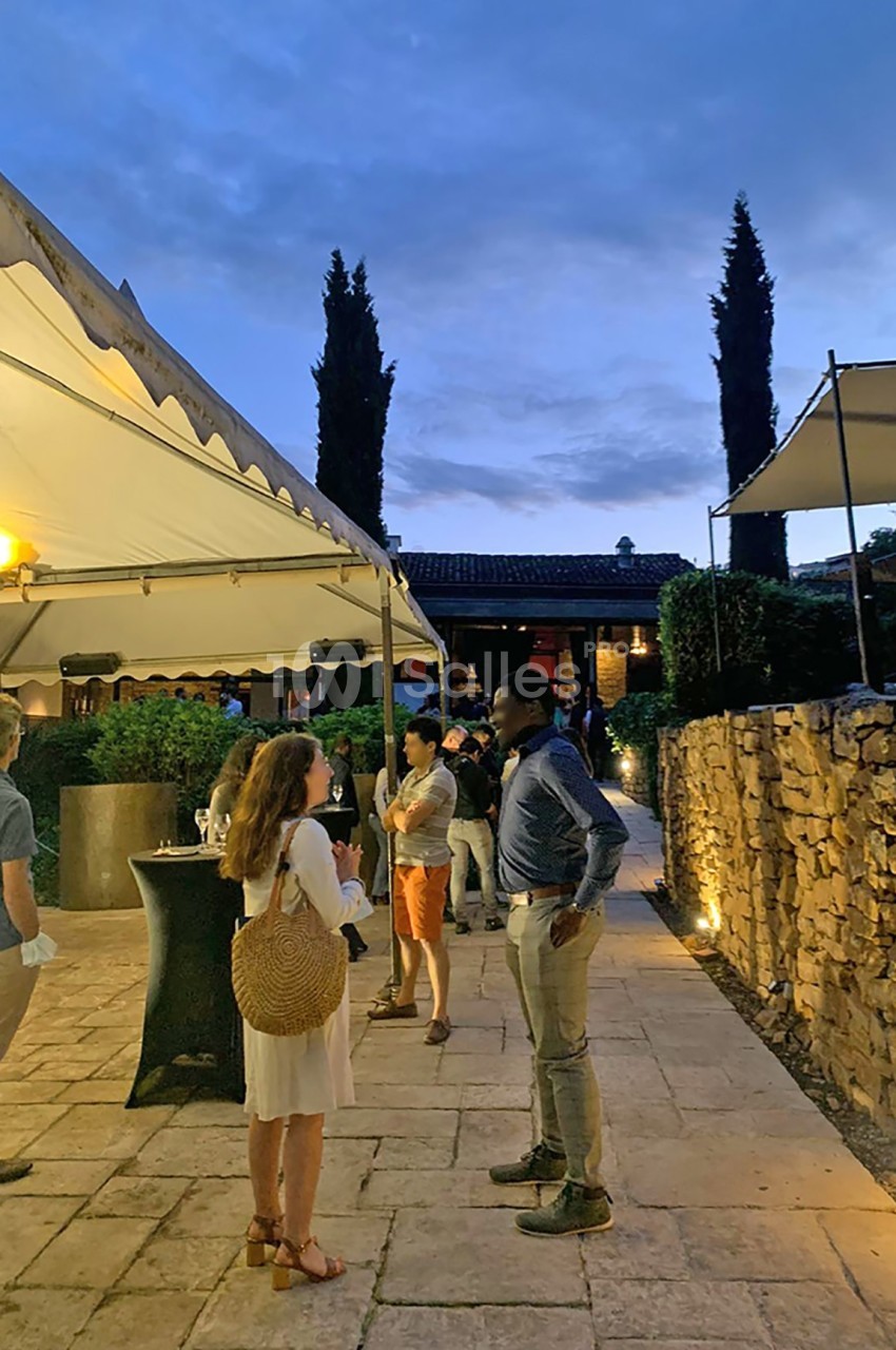 Personnes discutant dans une cour extérieure en soirée, éclairée par des lampes et entourée de murs en pierre.