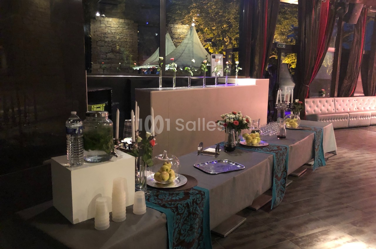 Tables décorées avec nappes et fleurs, disposées pour un buffet dans une salle éclairée avec des rideaux et des lumières…