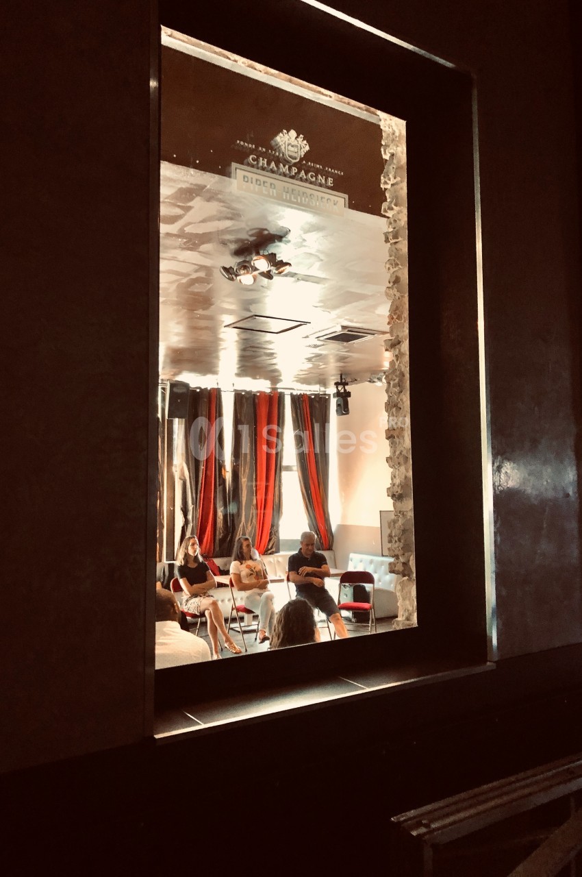 Vue à travers une fenêtre sur un groupe de personnes assises autour de tables dans une pièce aux rideaux rouges et noirs.