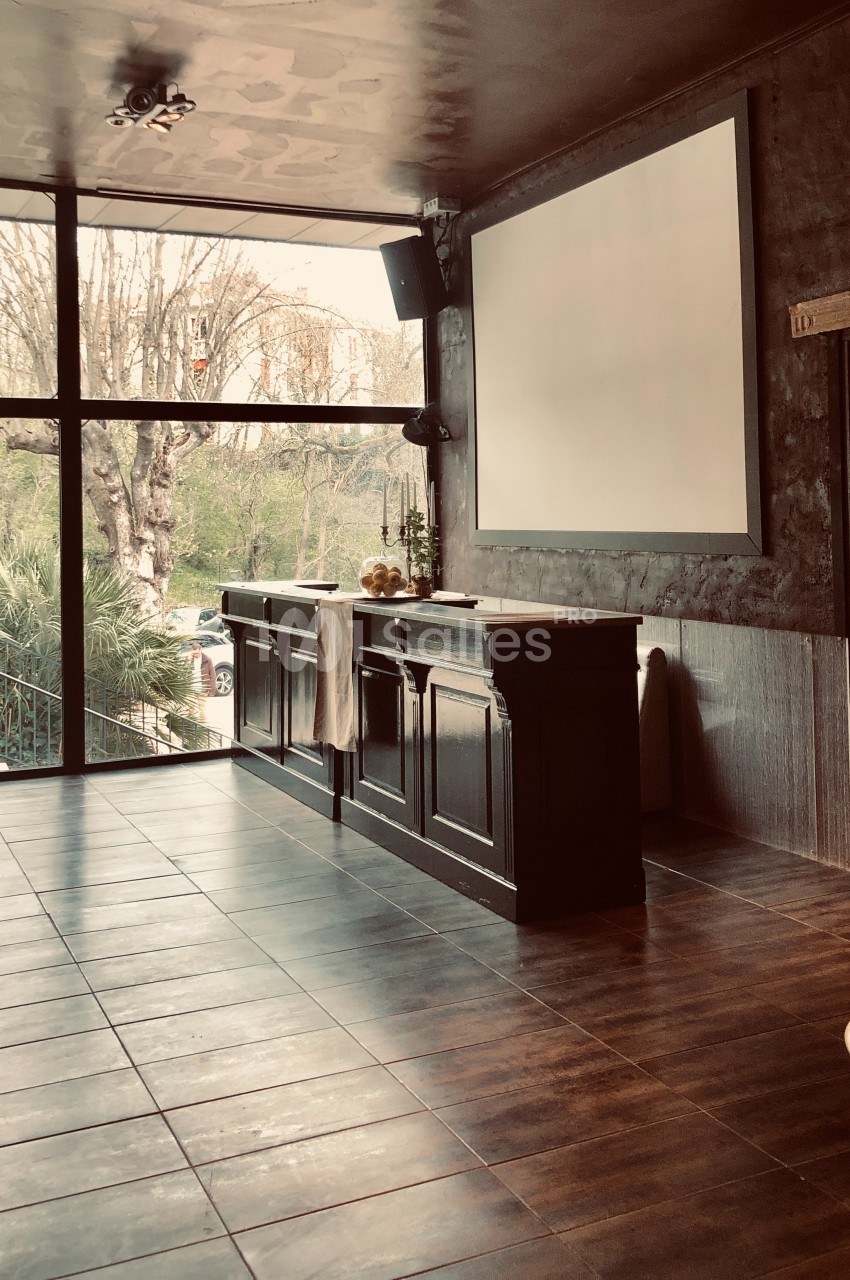 Salle lumineuse avec parquet sombre, grand écran blanc, bar en bois et vue sur des arbres à travers une baie vitrée.