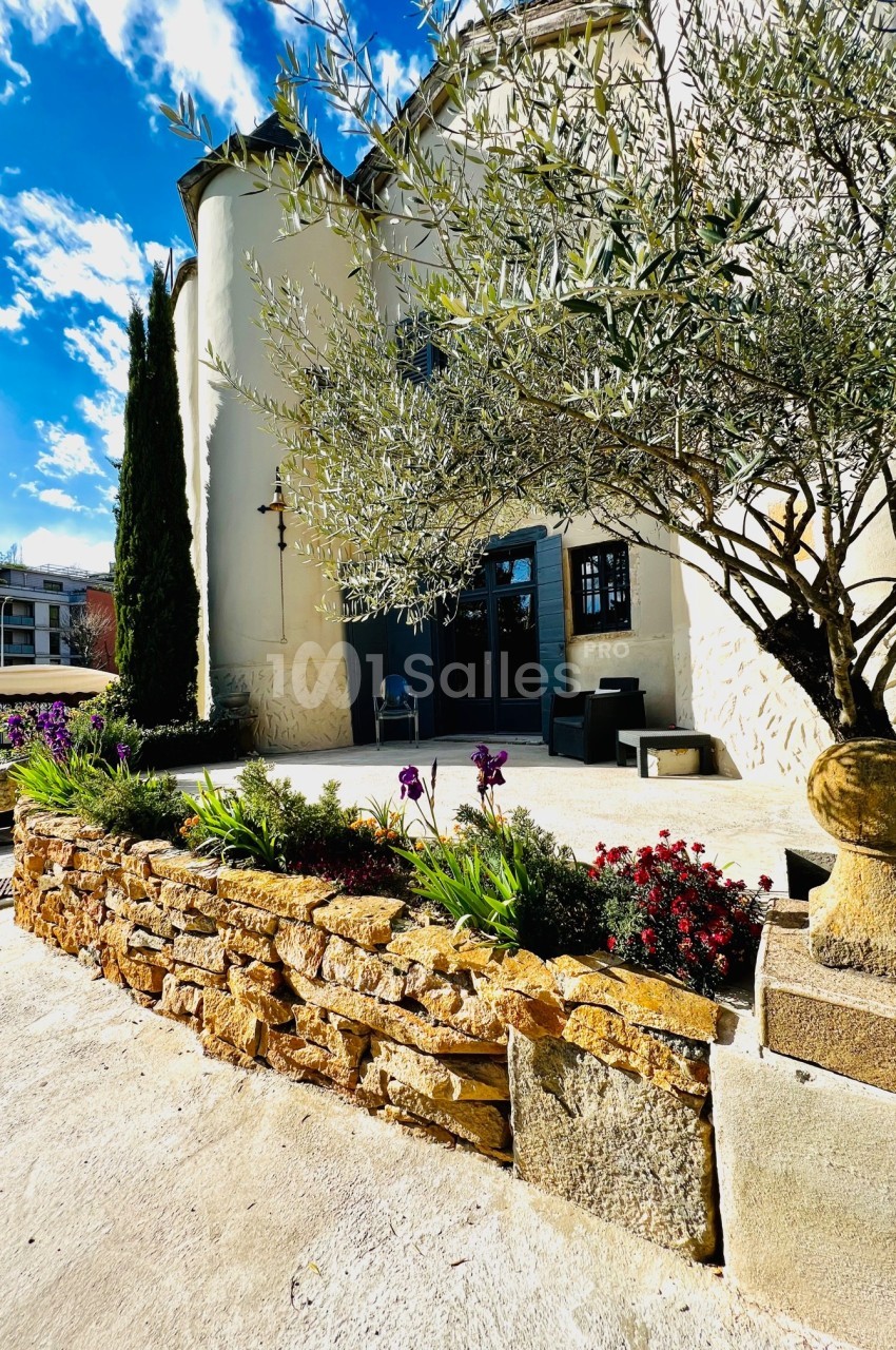 Façade d'un bâtiment blanc avec un olivier, des fleurs colorées et un muret en pierre sous un ciel ensoleillé.