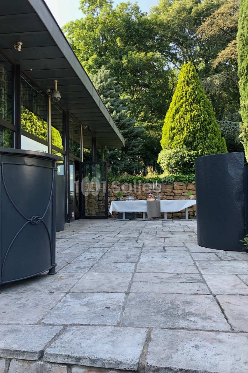 Terrasse en pierre avec table dressée, entourée de verdure et d'une façade vitrée moderne.