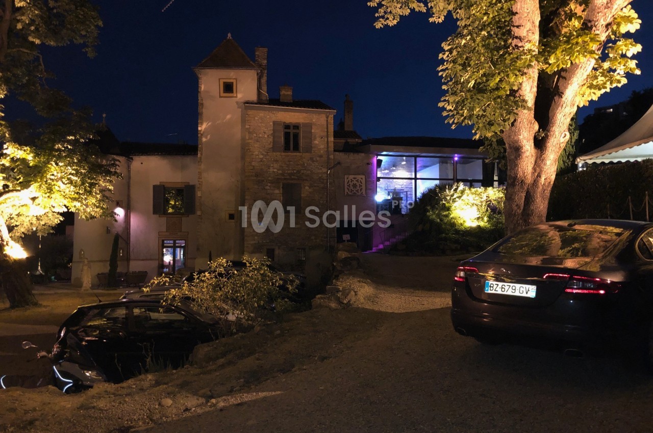 Vue nocturne d'un bâtiment ancien éclairé, entouré d'arbres, avec des voitures garées au premier plan.