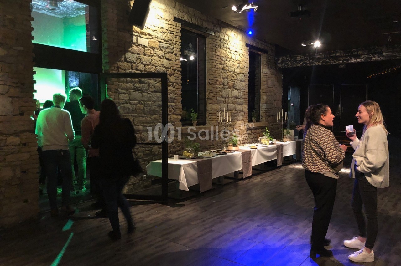 Des personnes discutent dans une salle avec murs en pierre, tandis qu’un groupe se dirige vers une zone éclairée en vert.