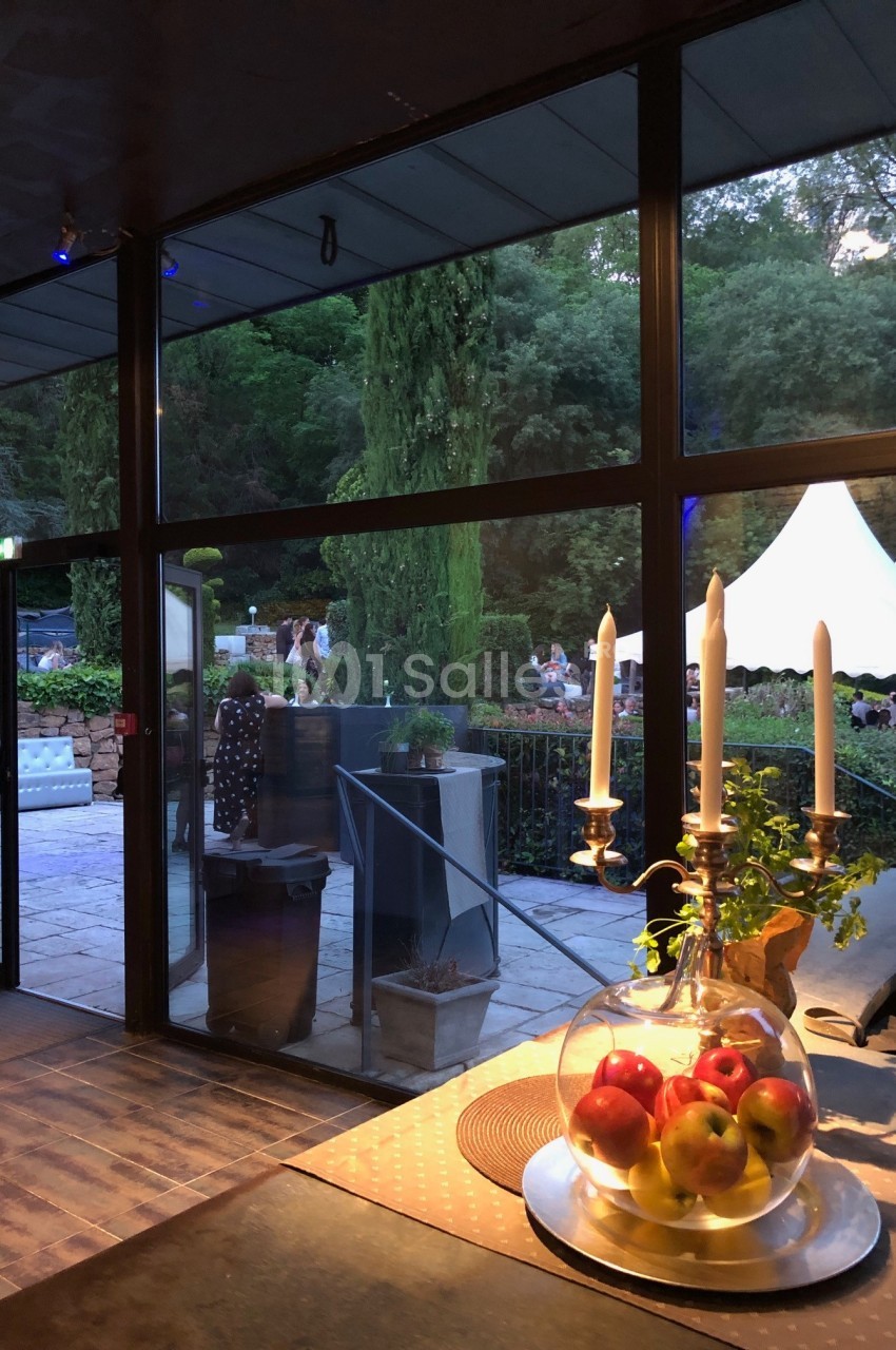 Vue sur un jardin avec des arbres et une tente blanche, depuis une pièce vitrée éclairée, avec un centre de table aux pommes.