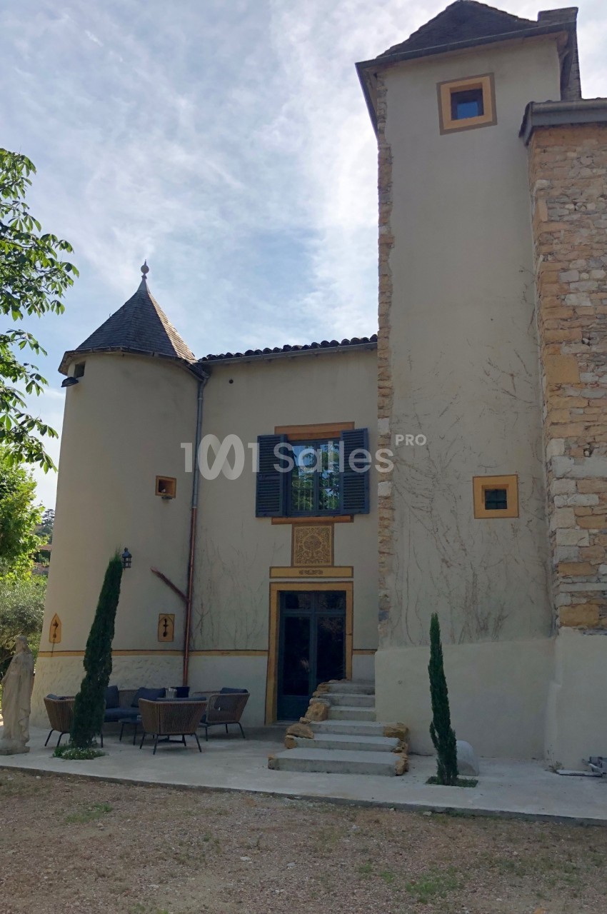 Façade d'une maison en pierre avec deux tours, fenêtres à volets et terrasse aménagée avec mobilier d'extérieur.