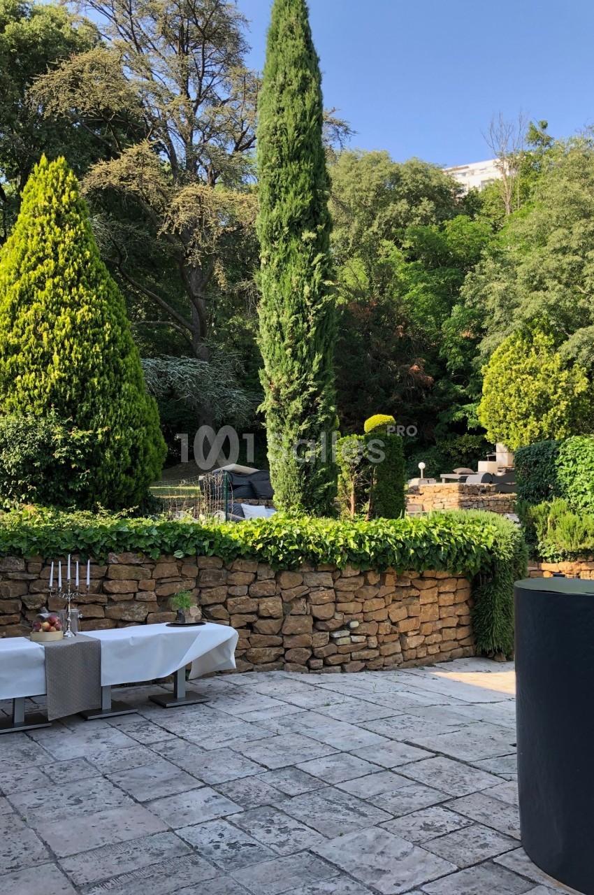 Terrasse en pierre avec tables dressées, bordée de murs en pierre et entourée de végétation dense et arbres.
