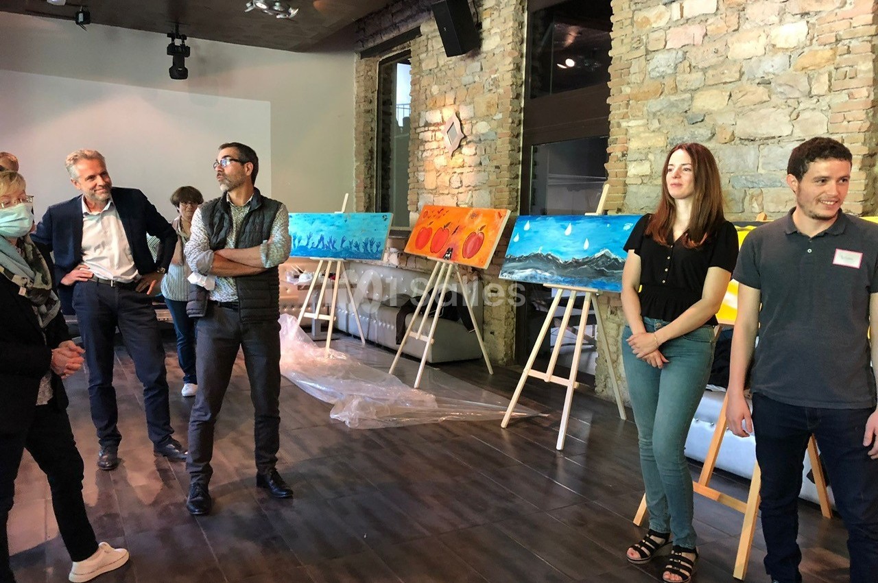 Des personnes debout dans une salle d'exposition avec des peintures colorées sur des chevalets devant un mur en pierre.