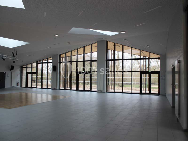 Intérieur d'une grande salle lumineuse avec des baies vitrées donnant sur un espace extérieur arboré.