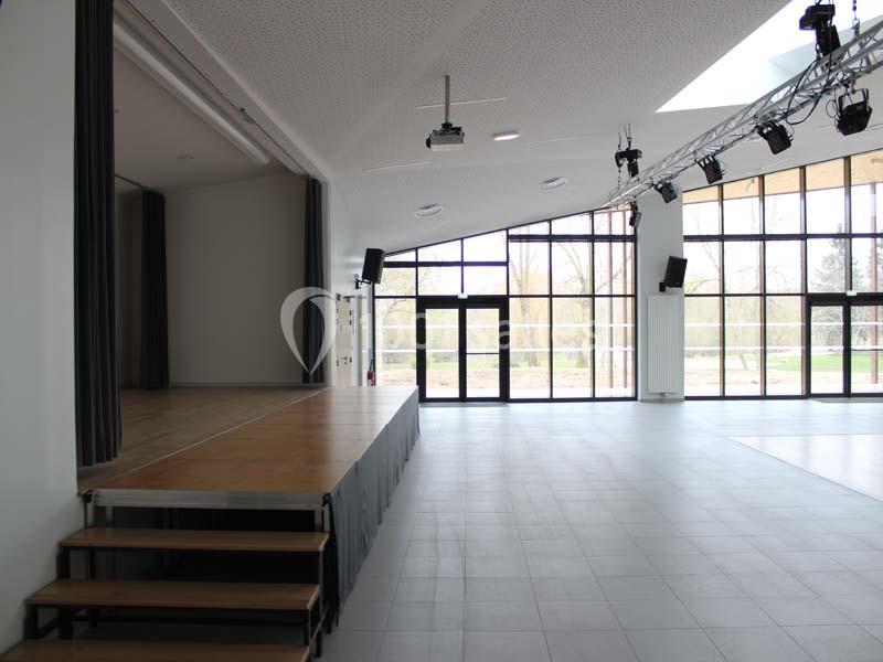 Salle lumineuse avec une scène en bois, des baies vitrées donnant sur l'extérieur et un éclairage suspendu.