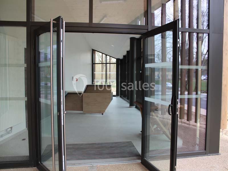 Entrée vitrée ouverte donnant sur un hall moderne avec comptoir en bois et vue sur l'extérieur arboré.