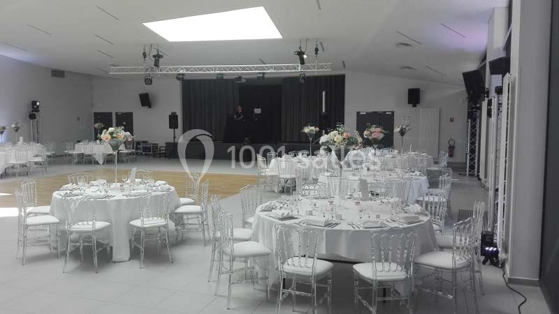 Salle de réception décorée avec des tables rondes, nappes blanches, chaises transparentes et centre de table floral.