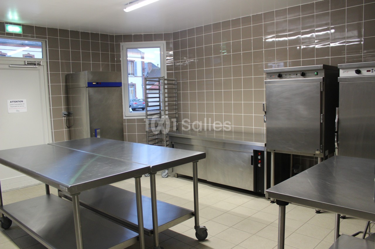 Cuisine professionnelle avec plans de travail en inox, étagères, fours et carrelage beige sur les murs.