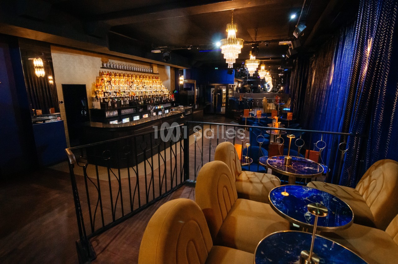 Intérieur d'un bar lounge avec comptoir éclairé, tables en marbre bleu et fauteuils rembourrés sous des lustres.