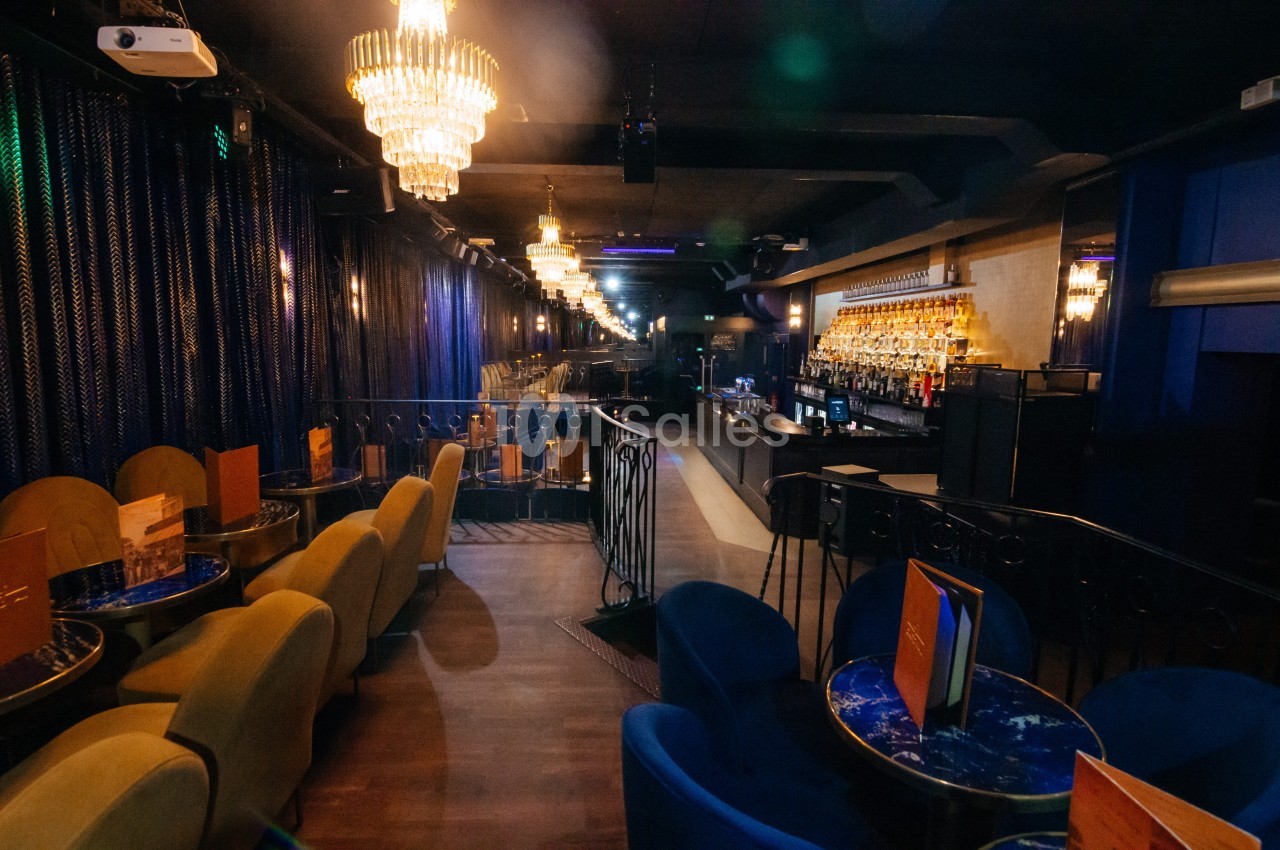 Salle de bar élégante avec lustres, fauteuils colorés, tables rondes et un comptoir illuminé en arrière-plan.