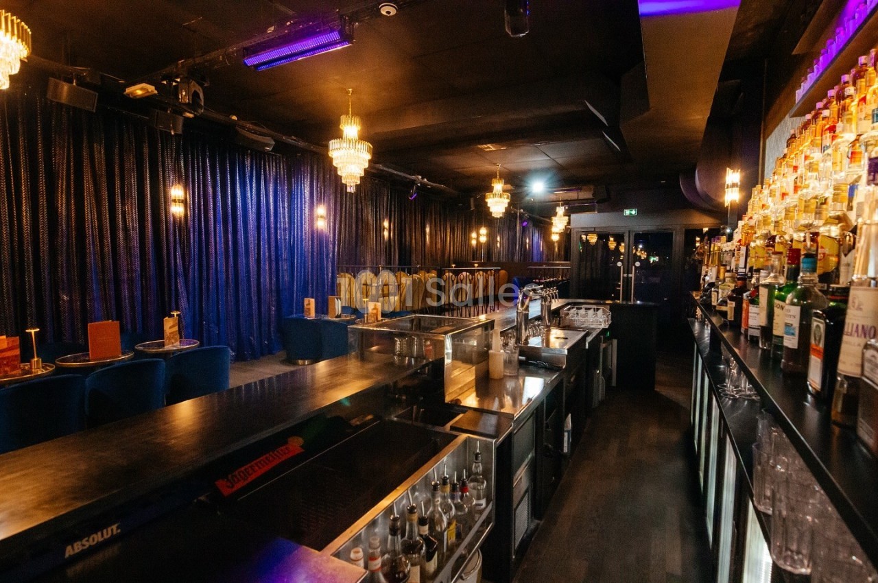 Salle de bar avec comptoir en bois, rangées de bouteilles, tables et chaises sous des lumières tamisées.