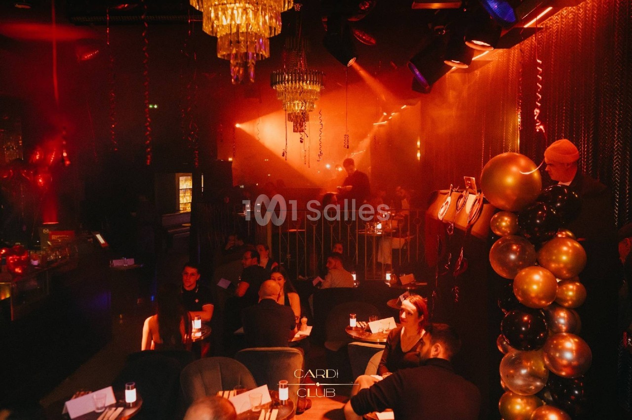Salle de club avec éclairage tamisé, décorations de ballons et invités assis autour de tables.