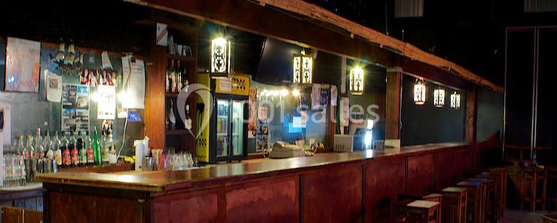 Intérieur d'un bar avec un long comptoir en bois, des tabourets alignés et des étagères remplies de bouteilles.