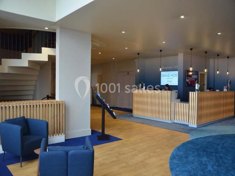 Réception d'un hôtel avec comptoir en bois, fauteuils bleus, escalier en colimaçon et éclairage moderne.