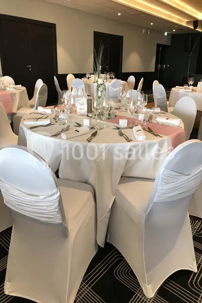 Salle de réception avec tables rondes dressées, nappes blanches, chaises houssées et décoration florale centrale.
