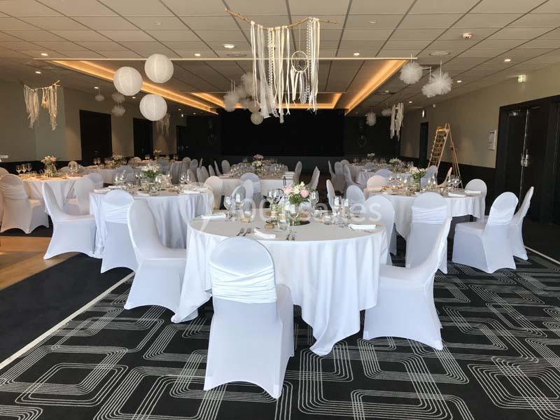 Salle de réception décorée avec des tables rondes, nappes blanches, chaises houssées et suspensions en papier.