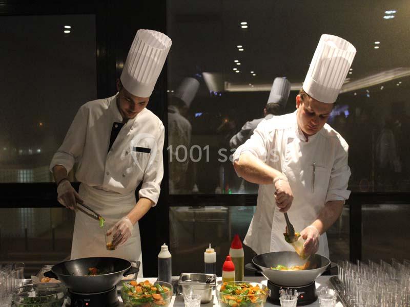 Deux chefs en tenue blanche préparent des plats dans des woks sur un comptoir, entourés de condiments.