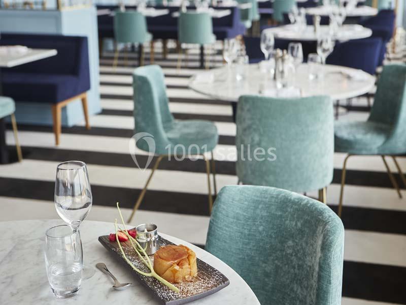 Assiette avec un dessert et des couverts sur une table en marbre dans un restaurant aux chaises bleues et sol rayé.