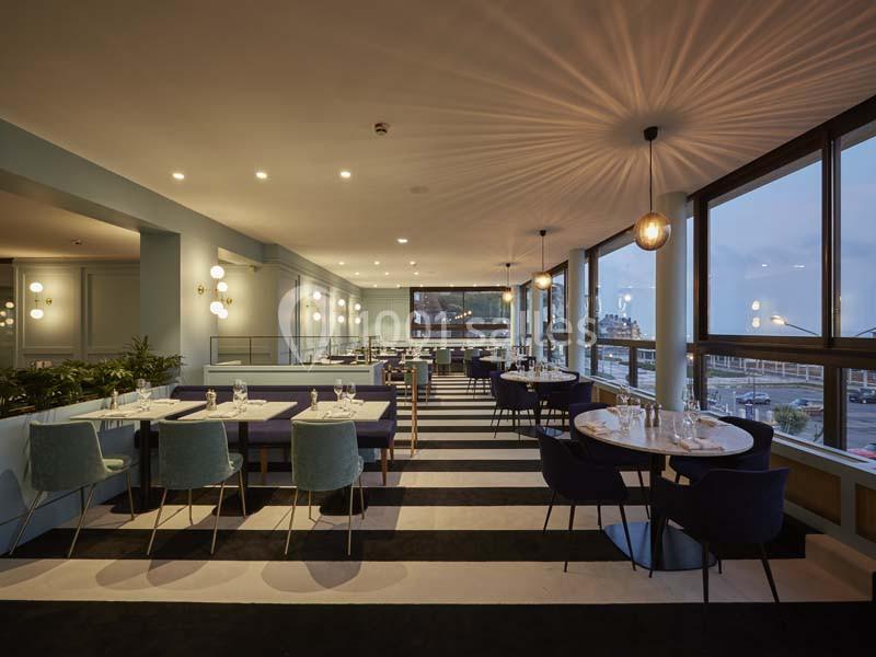 Salle de restaurant moderne avec tables dressées, chaises colorées et grandes baies vitrées laissant entrer la lumière…