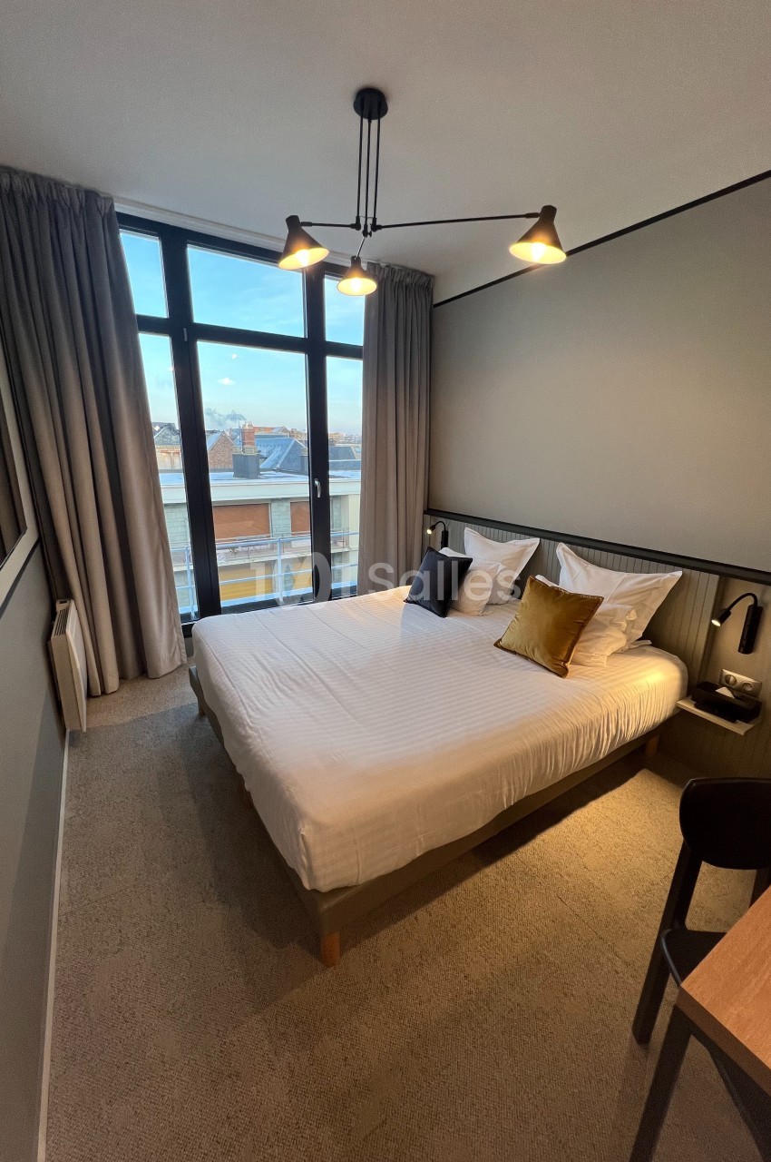 Chambre moderne avec lit double, grandes fenêtres donnant sur des bâtiments, éclairage suspendu et décor sobre.