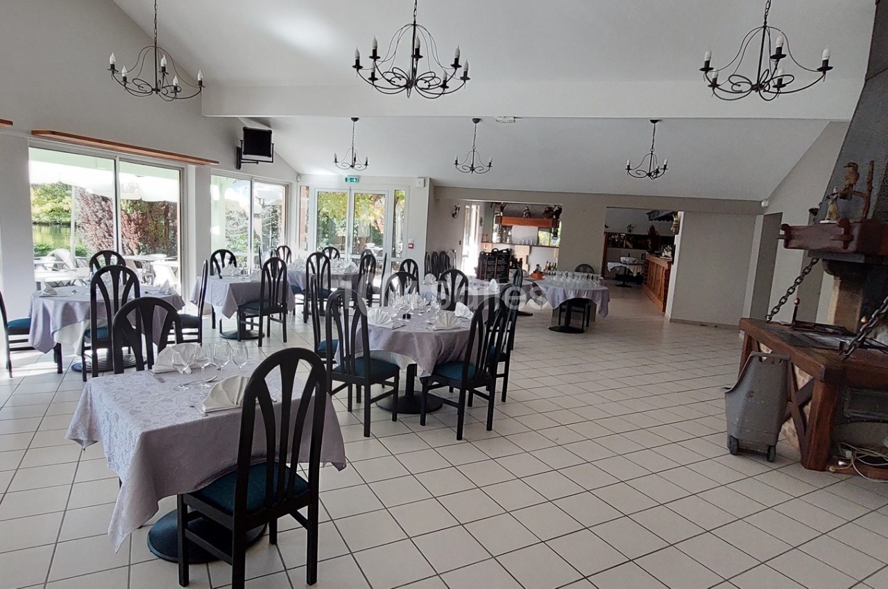 Salle de restaurant lumineuse avec tables dressées, chaises noires et cheminée décorative dans un cadre spacieux.