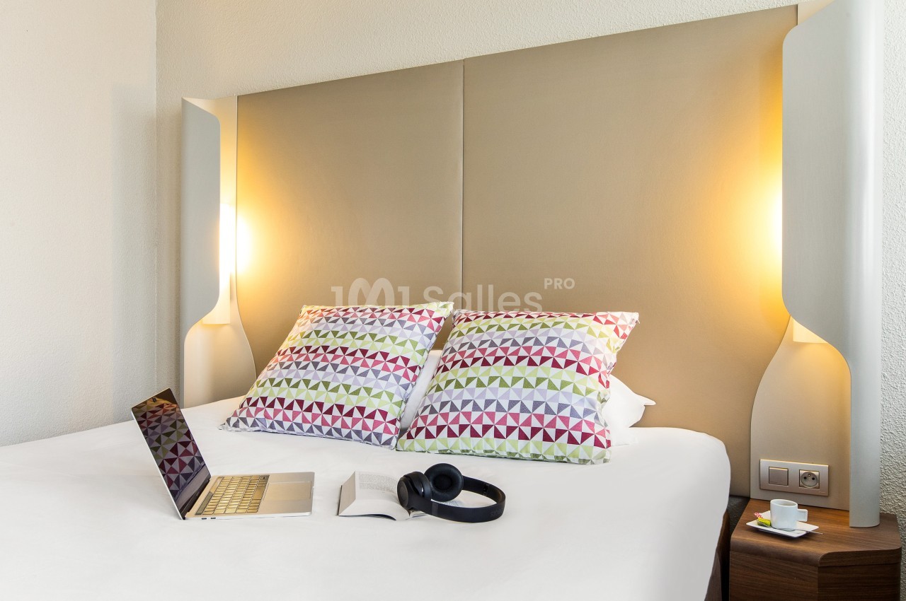 Lit double avec tête de lit beige, oreillers à motifs géométriques colorés, ordinateur portable et casque audio posés dessus.