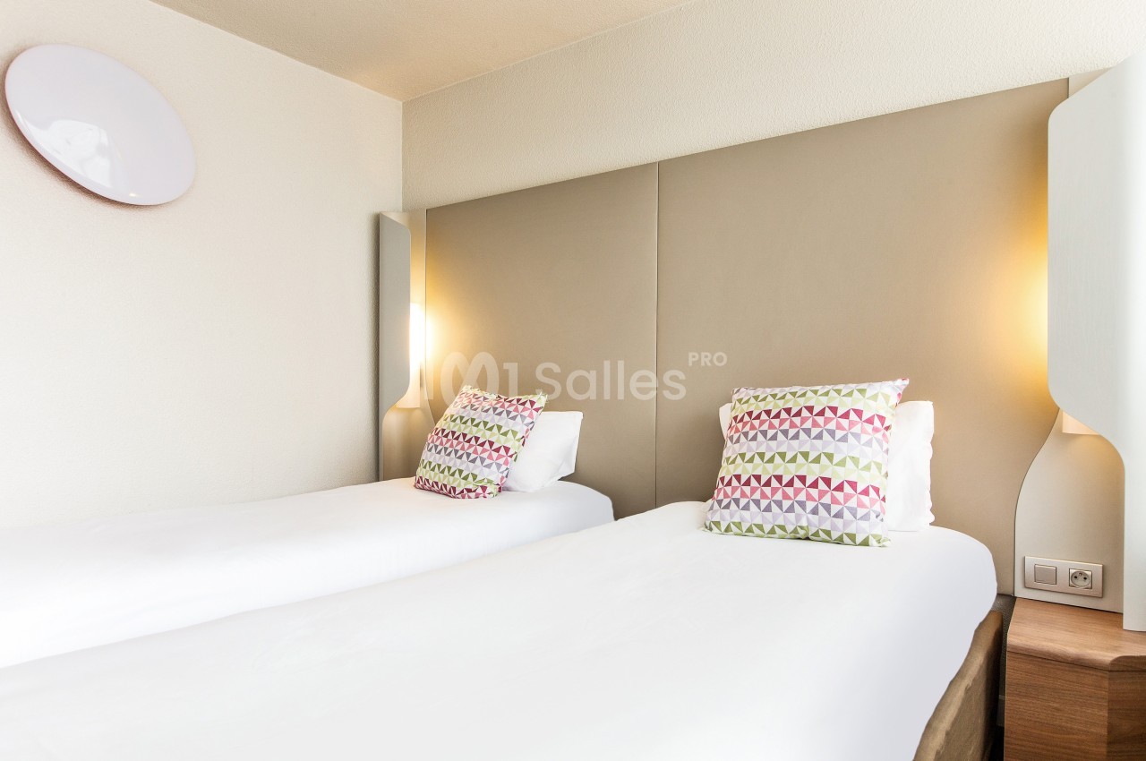 Deux lits simples avec des coussins colorés, éclairés par des appliques murales dans une chambre lumineuse.
