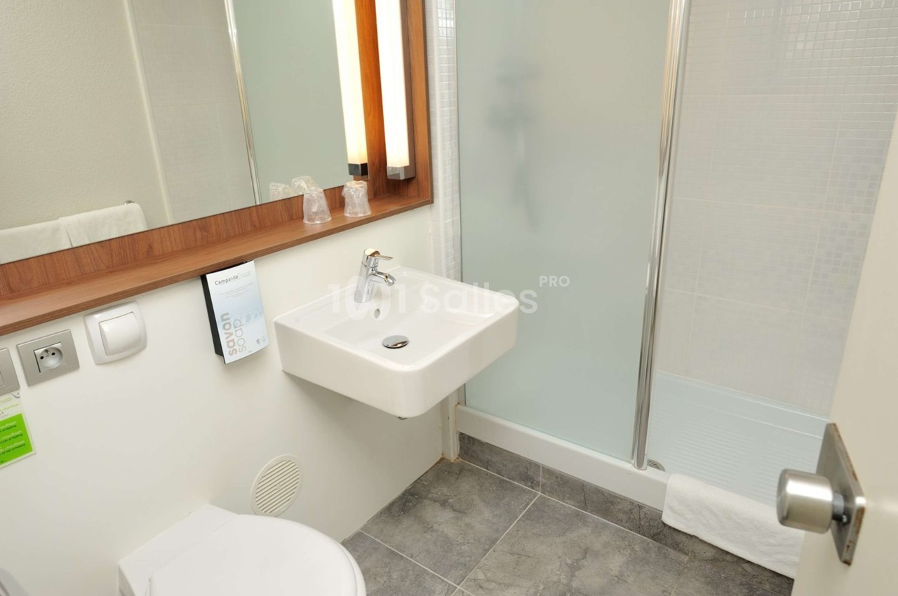 Salle de bain avec lavabo mural, miroir, douche vitrée et sol en carrelage gris.