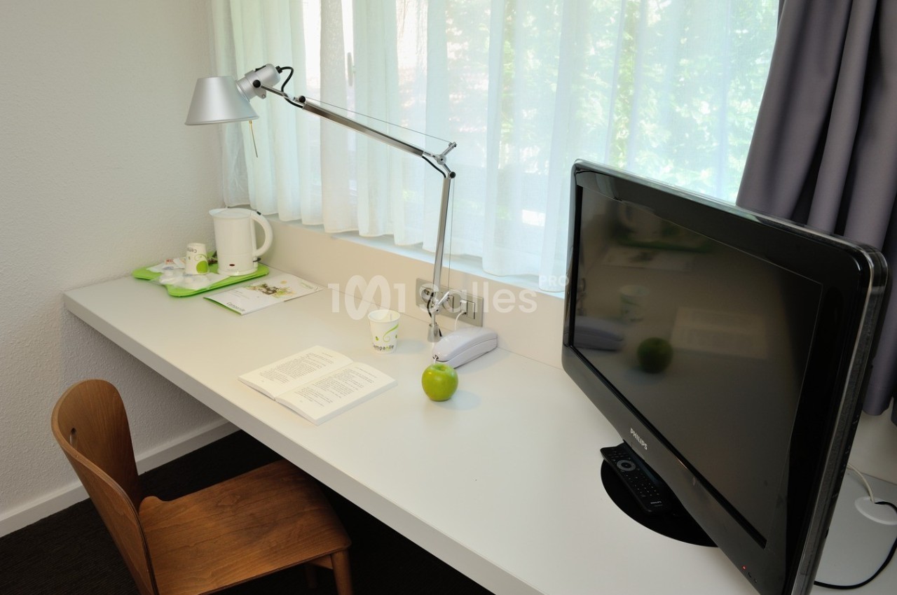 Bureau avec lampe articulée, télévision, chaise en bois, livre ouvert, pomme verte et plateau avec bouilloire.