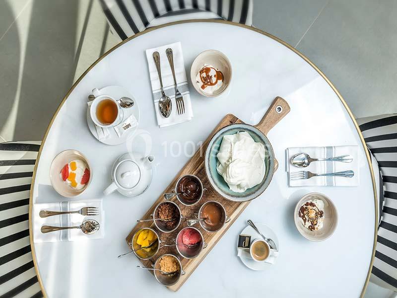 Table vue de dessus avec un plateau de desserts variés, une théière, des tasses et des couverts disposés autour.