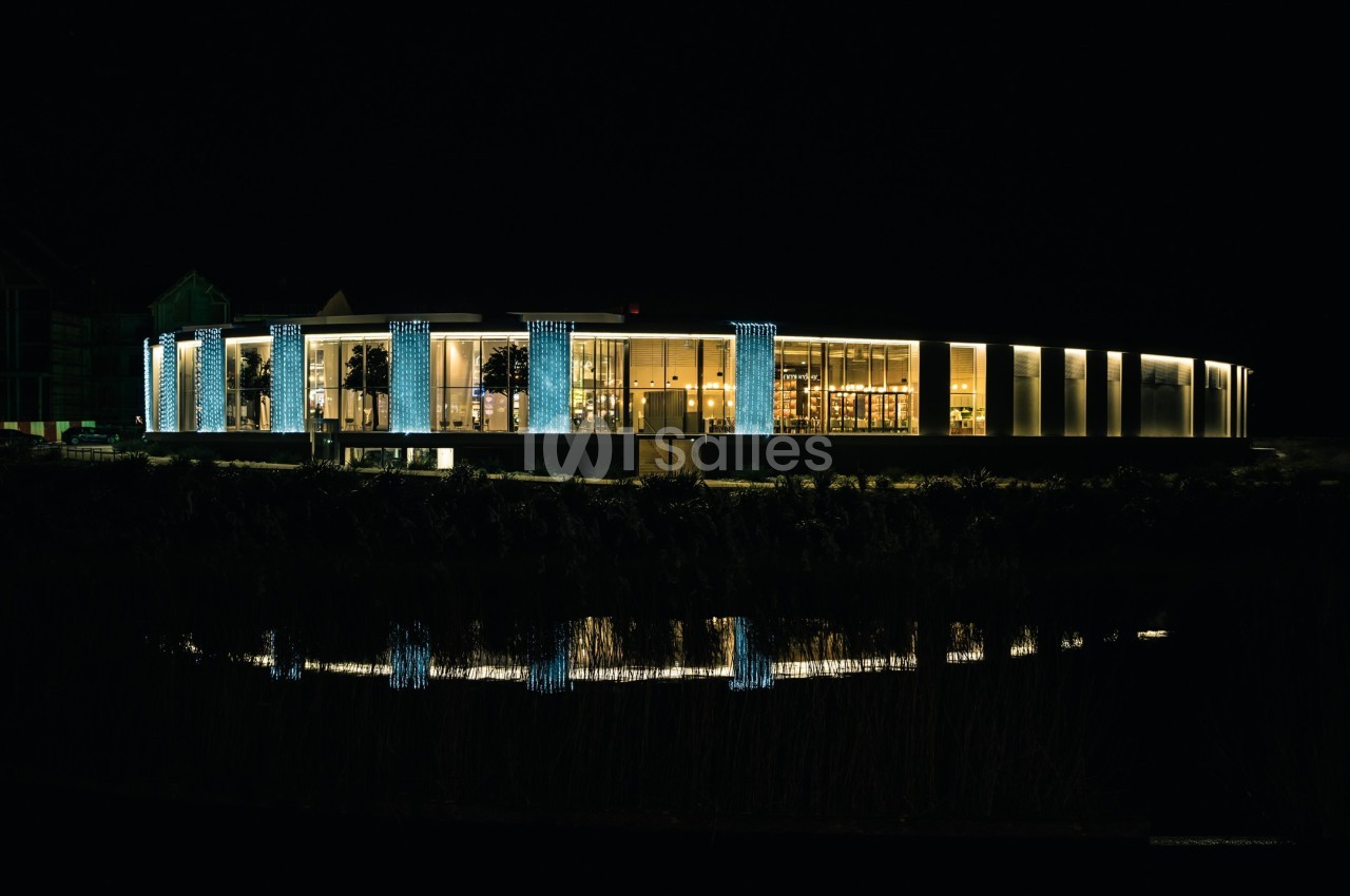 Bâtiment moderne éclairé la nuit, avec des colonnes lumineuses et son reflet visible dans l'eau au premier plan.