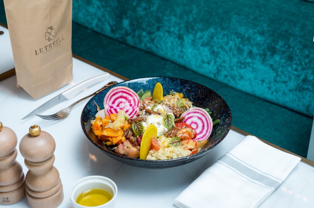Bol de salade colorée avec légumes, betteraves, couscous et sauce, servi sur une table avec couverts et huile d'olive.