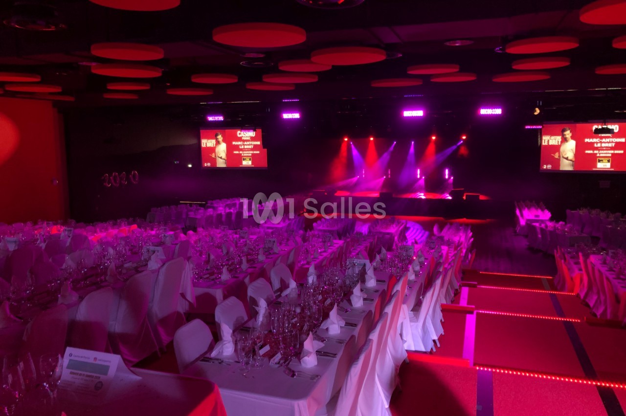 Salle de réception éclairée en rouge avec tables dressées et une scène illuminée en arrière-plan.