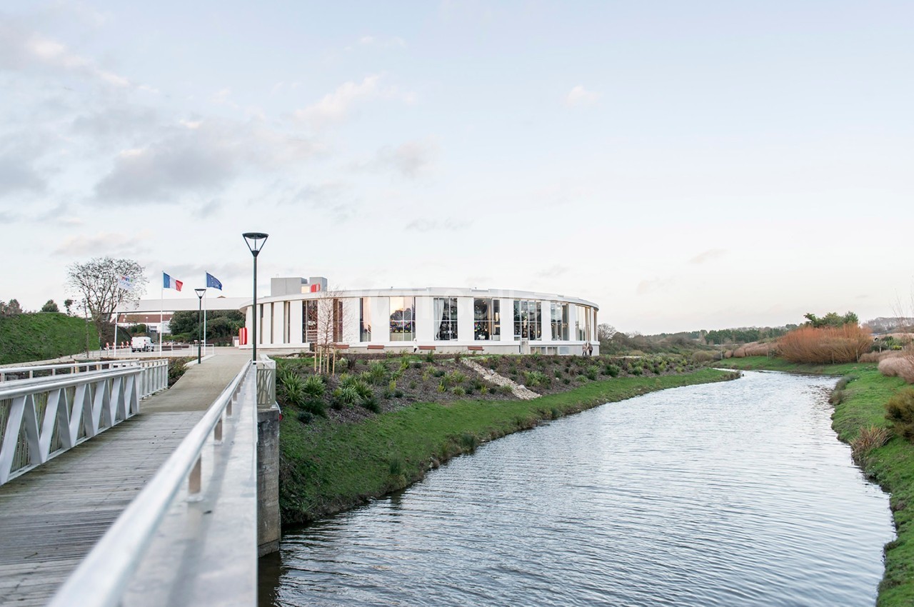 Bâtiment moderne en bord de rivière, entouré de verdure, avec un pont piétonnier au premier plan.
