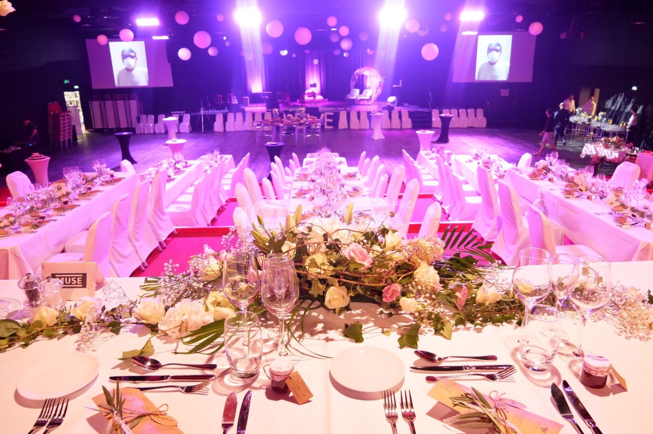 Salle de réception décorée avec des tables dressées, des fleurs et un éclairage rose lumineux.