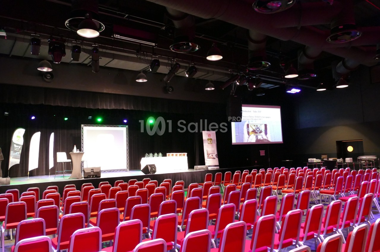 Salle de conférence avec rangées de chaises rouges, une scène éclairée et un écran de projection allumé.