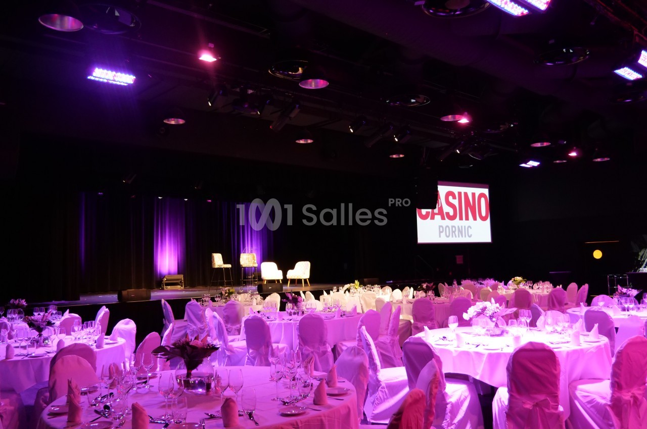 Salle de réception éclairée en rose avec tables dressées et scène équipée de fauteuils, au Casino de Pornic.