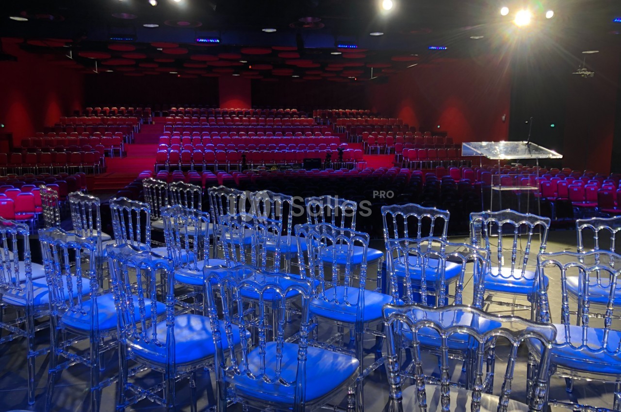 Salle de conférence avec des rangées de chaises transparentes et une scène éclairée par des projecteurs.