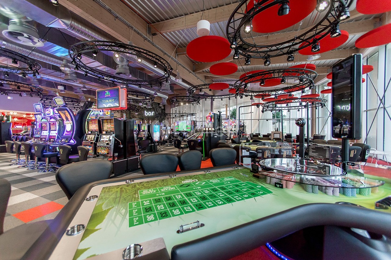 Salle de casino moderne avec tables de jeu, machines à sous et éclairage décoratif rouge au plafond.