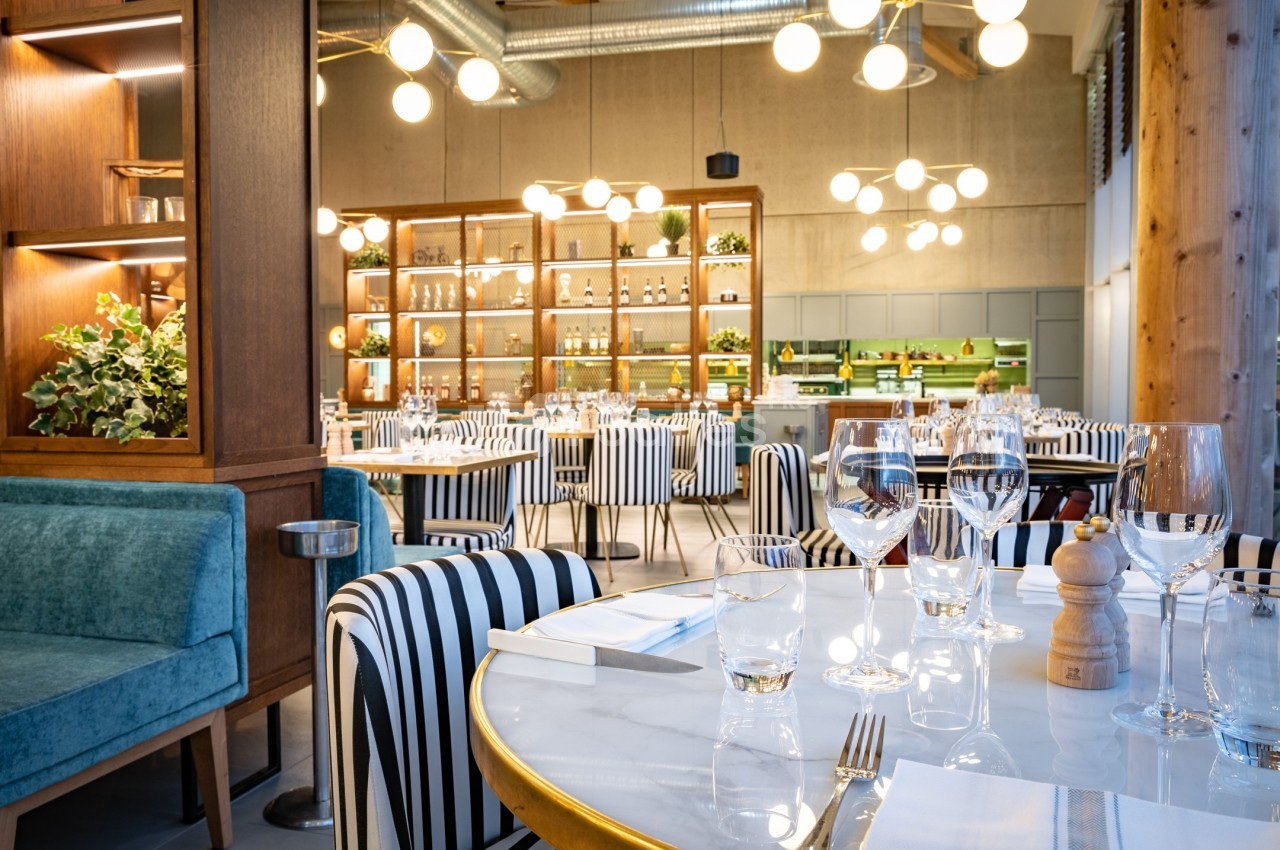 Salle de restaurant moderne avec tables dressées, chaises à rayures, étagères décoratives et éclairage suspendu.