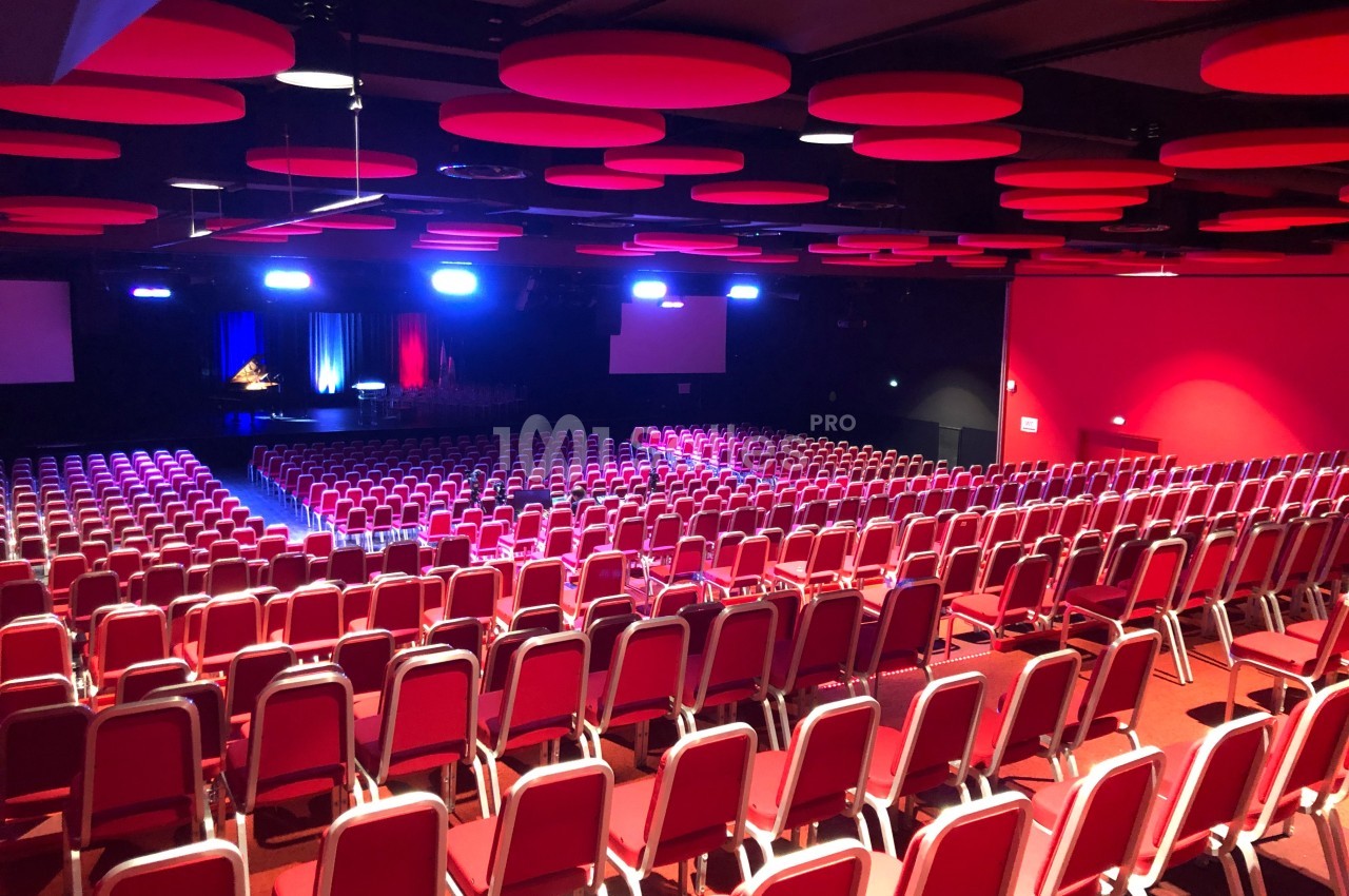 Salle de conférence avec rangées de chaises rouges, éclairage coloré et scène équipée d'écrans et rideaux.