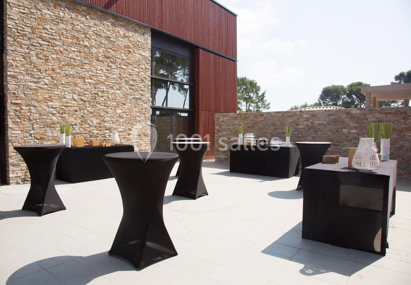 Terrasse aménagée avec des tables hautes et buffets noirs, entourée de murs en pierre et d'un bâtiment moderne.