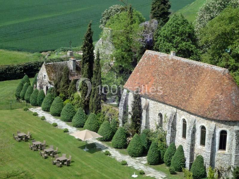 Vue aérienne d'un jardin avec pelouse, tables, chaises, et bâtiments en pierre entourés de haies et d'arbres.